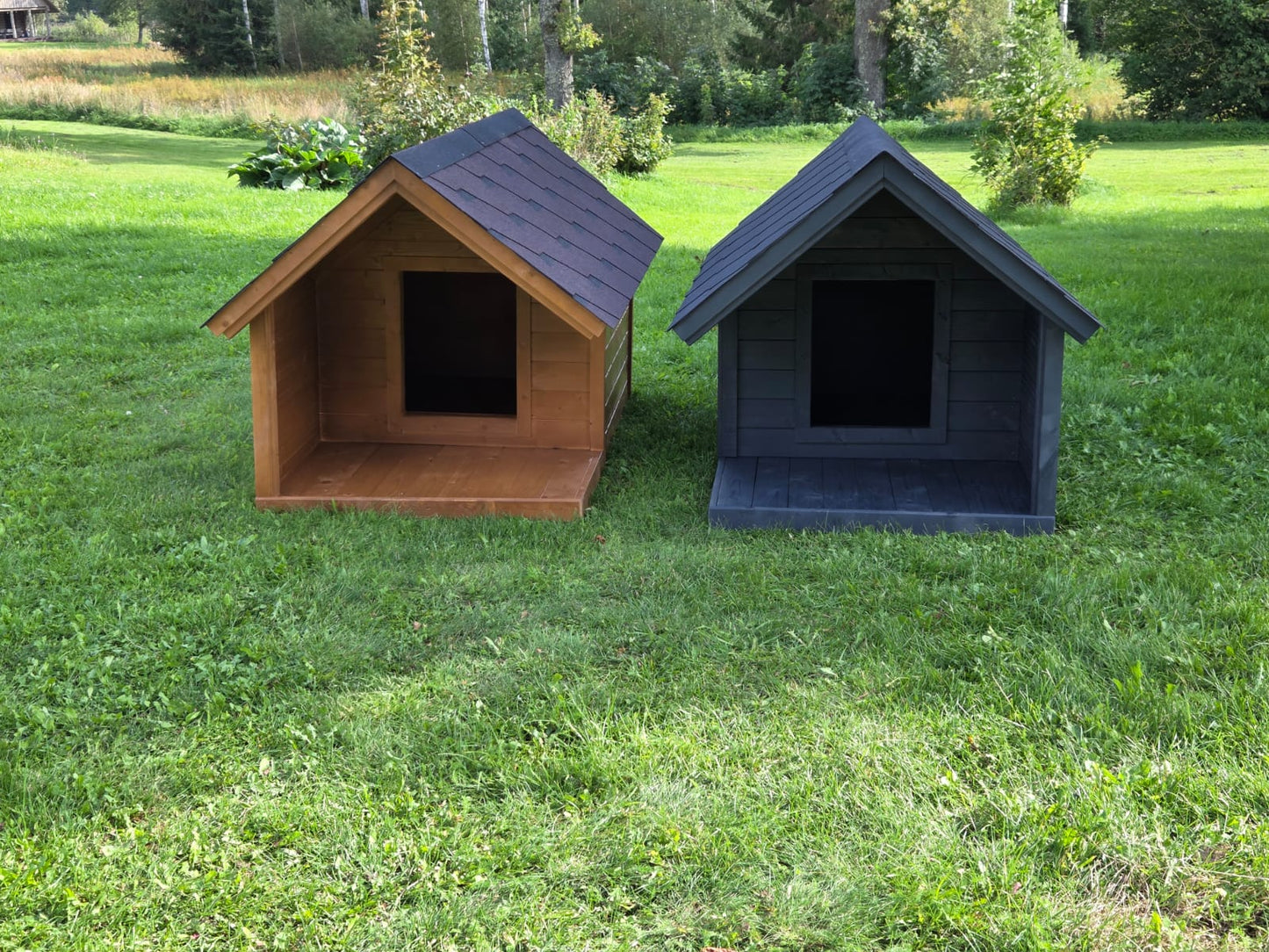Two dog houses, one brown and one black, on a grassy field with trees in the background.