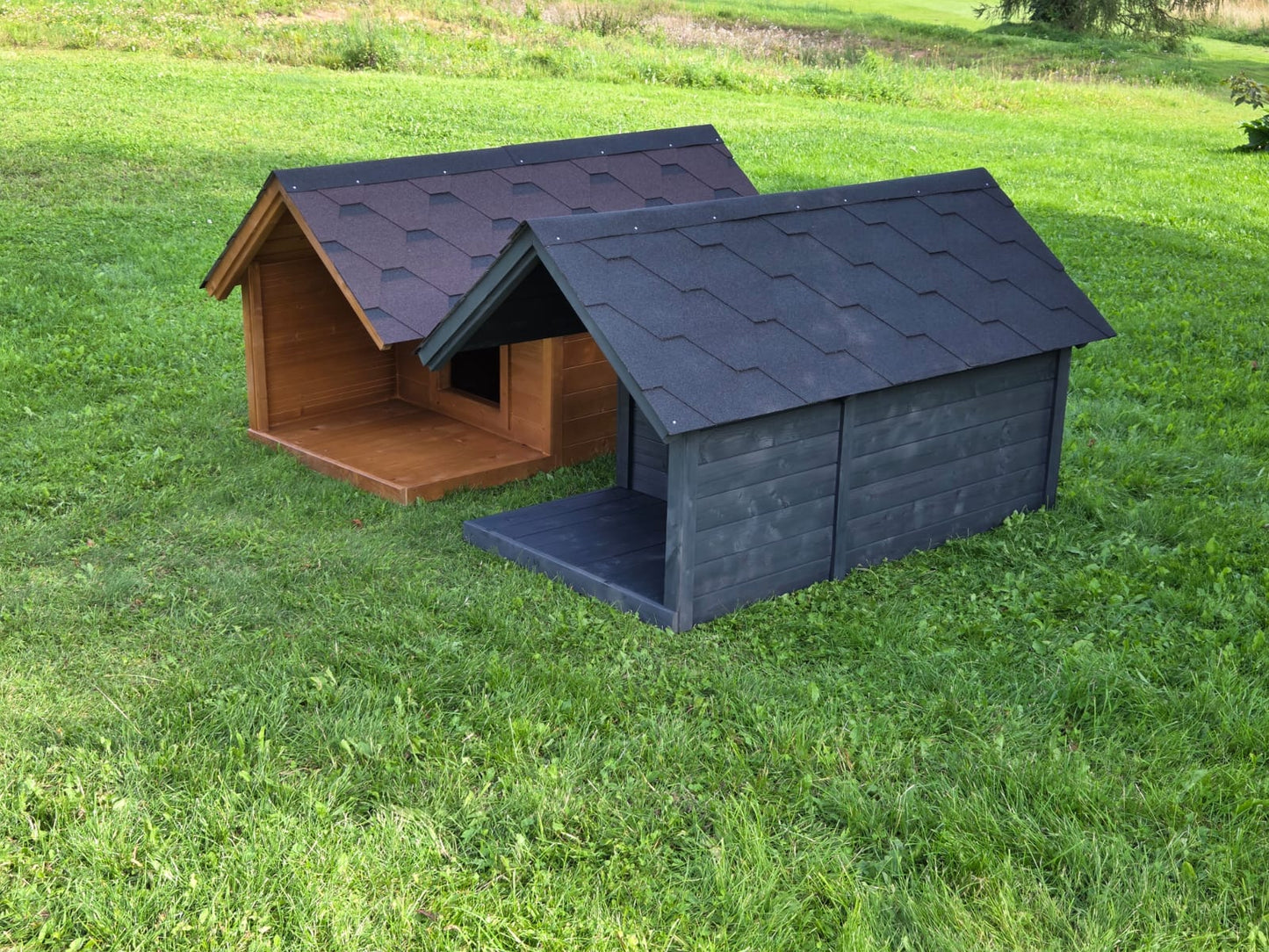 Two dog houses, one brown and one gray, on a grassy field.