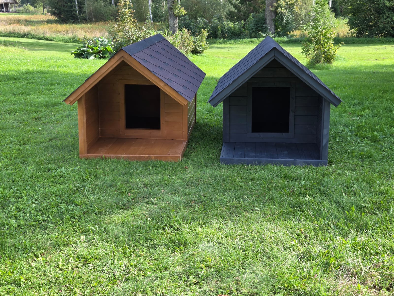 Two dog houses, one brown and one black, on a grassy field with trees in the background.